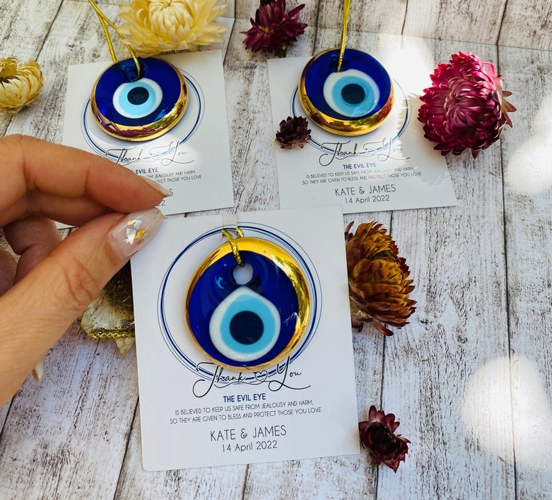 K&ouml;nnte beinhalten: Blauer und wei&szlig;er b&ouml;ser Blick-Anh&auml;nger mit goldfarbenem Rand, der an einer goldenen Schnur h&auml;ngt. Der Anh&auml;nger befindet sich auf einer wei&szlig;en Karte mit dem Text "The Evil Eye is believed to keep us safe from jealousy and harm, so they are given to bless and protect those you love. Kate & James 14 April 2022."