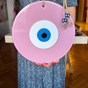 K&ouml;nnte beinhalten: Ein gro&szlig;er, runder, rosa Wandbehang mit einem blau-wei&szlig;en Augendesign. Das dekorative St&uuml;ck ist an einem Naturseil aufgeh&auml;ngt und verf&uuml;gt &uuml;ber zus&auml;tzliche blaue Augenanh&auml;nger. Das augenf&ouml;rmige Design ist ein beliebtes Schutzsymbol.