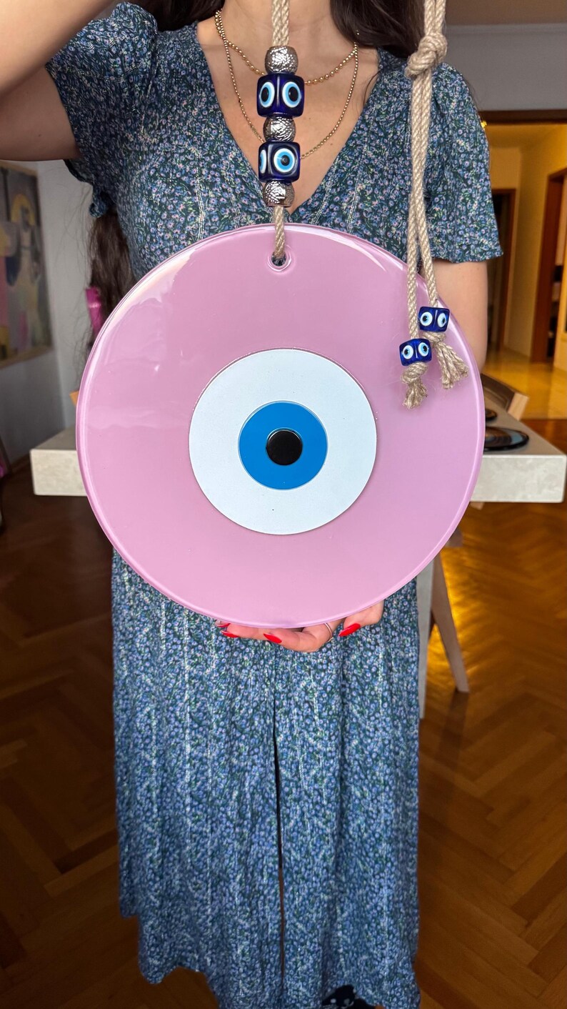 K&ouml;nnte beinhalten: Ein gro&szlig;er, runder, rosa Wandbehang mit einem blau-wei&szlig;en Augendesign. Das dekorative St&uuml;ck ist an einem Naturseil aufgeh&auml;ngt und verf&uuml;gt &uuml;ber zus&auml;tzliche blaue Augenanh&auml;nger. Das augenf&ouml;rmige Design ist ein beliebtes Schutzsymbol.