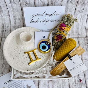 Può includere: Una scatola regalo bianca piena di oggetti per un rituale spirituale. La scatola contiene un piatto in ceramica bianca con un simbolo zodiacale Gemelli in oro, un ciondolo di occhio blu e bianco, un mazzo di salvia secca, un piccolo bastoncino di incenso Palo Santo in legno e una candela di cera d'api gialla. La scatola è decorata con carta bianca triturata. Il testo sulla carta dice "güzel şeyleri hak ediyorsun", che si traduce in "Ti meriti cose belle".