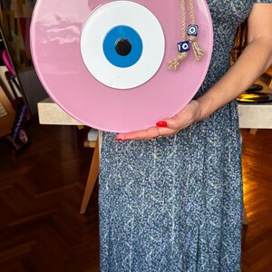 K&ouml;nnte beinhalten: Ein gro&szlig;er, runder, rosa Evil Eye Wandbehang mit blauem und wei&szlig;em Augendesign. Das dekorative St&uuml;ck ist an einem Seil aufgeh&auml;ngt und hat oben eine blaue Perle. Ein einzigartiger Einrichtungsgegenstand.