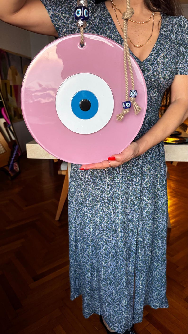 K&ouml;nnte beinhalten: Ein gro&szlig;er, runder, rosa Evil Eye Wandbehang mit blauem und wei&szlig;em Augendesign. Das dekorative St&uuml;ck ist an einem Seil aufgeh&auml;ngt und hat oben eine blaue Perle. Ein einzigartiger Einrichtungsgegenstand.