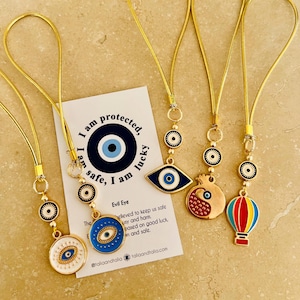 May include: Five gold chain phone charms with blue and white evil eye designs. The charms are attached to a card with the text "I am protected, I am safe, I am lucky." and "Evil Eye" with a description of the evil eye.