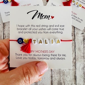 May include: Red string bracelet with a blue evil eye charm, a gold heart charm, and white beads spelling out the name "TALIA". The bracelet is tied around a white card with the text "I hope with this red string and evil eye bracelet all your wishes will come true and protected you from everything." and "Happy Mother's Day Thank you for always being there for me, Love you today, tomorrow and always."