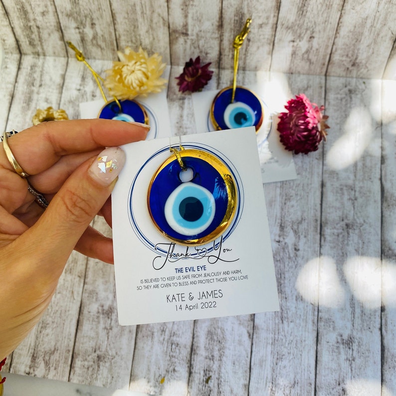 K&ouml;nnte beinhalten: Blauer und wei&szlig;er b&ouml;ser Blick-Anh&auml;nger mit goldfarbenem Rand, der an einer goldenen Schnur h&auml;ngt. Der Anh&auml;nger befindet sich auf einer wei&szlig;en Karte mit dem Text "Danke" und "Der b&ouml;se Blick soll uns vor Neid und Schaden sch&uuml;tzen, daher wird er gegeben, um diejenigen zu segnen und zu sch&uuml;tzen, die du liebst Kate & James 14. April 2022."