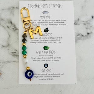 Op de afbeelding: Een goudkleurige sleutelhanger met de letter "M" en een kralenketting van groene, witte en gouden kralen, en een evil eye bedel. De bedel is bevestigd aan een kaart met de tekst "FOR YOUR NEXT CHAPTER".