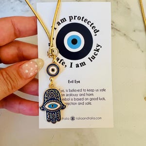 Puede incluir: Collar dorado con un colgante de ojo turco y mano de Fátima. El colgante es azul marino con detalles blancos y dorados. La tarjeta detrás dice: "Estoy protegido, seguro, tengo suerte."