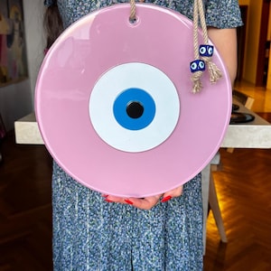 This image shows a large handcrafted pink evil eye wall hanging being held for display. The piece features glossy glass layers in pink, white, blue, and black, accented with gold beads and a traditional blue cube bead on natural rope. This stylish Turkish Nazar ornament is designed for home protection, positive energy, and modern boho decor.
