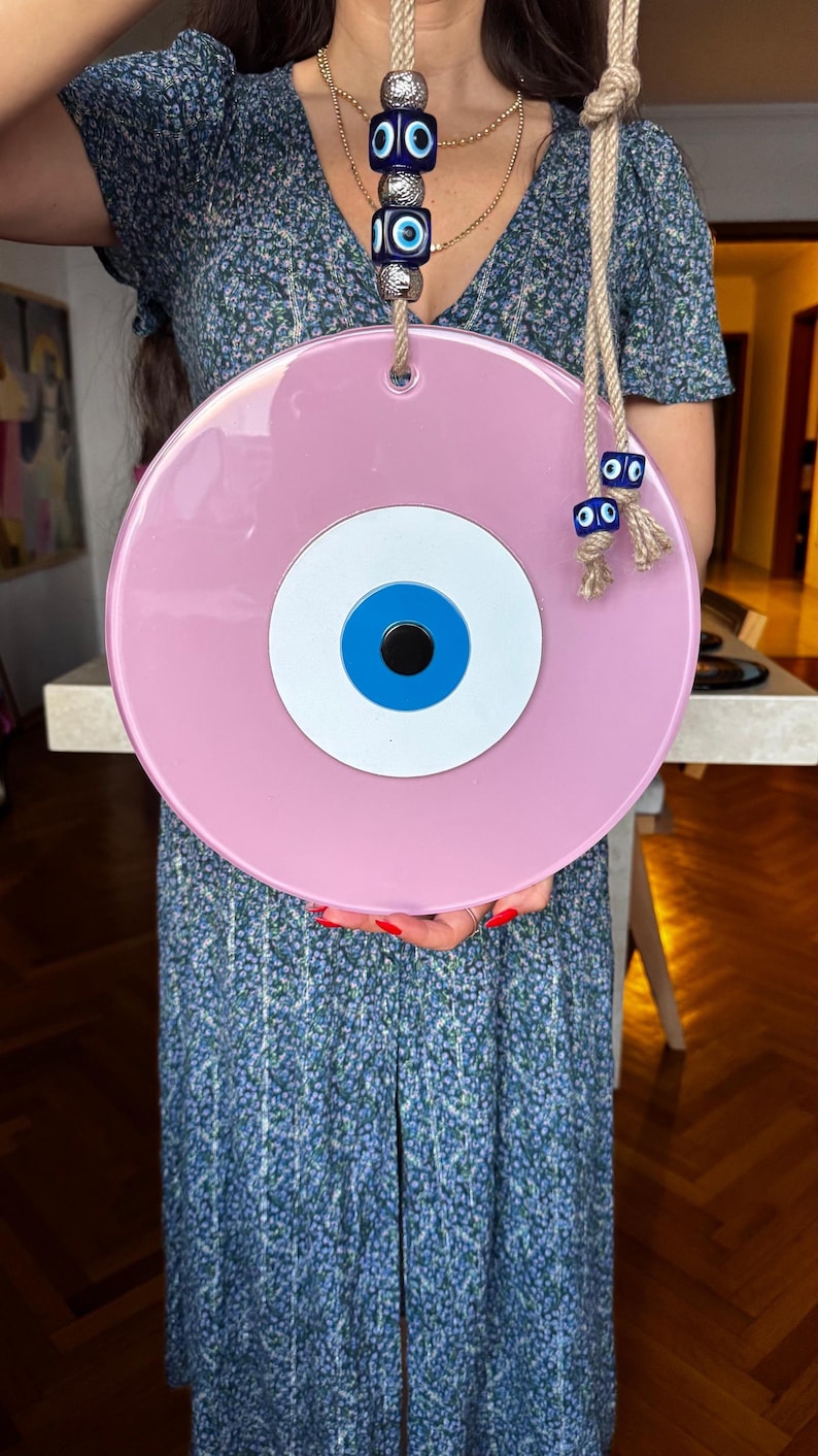 This image shows a large handcrafted pink evil eye wall hanging being held for display. The piece features glossy glass layers in pink, white, blue, and black, accented with gold beads and a traditional blue cube bead on natural rope. This stylish Turkish Nazar ornament is designed for home protection, positive energy, and modern boho decor.