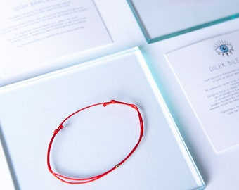 May include: Red string bracelet with a gold bead. The bracelet is on a white surface with a card that says "Dilek Bilezik" in black text.