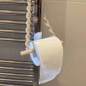 May include: A white toilet roll is hanging from a wooden dowel rod with a beige macrame rope. The rope is attached to a chrome towel rack.