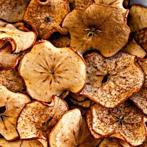 May include: A close-up shot of dried apple slices. The slices are a warm, golden-brown colour with darker brown speckles and edges. The core of each slice is visible, with a star-shaped seed pattern. The slices vary in shape and size.