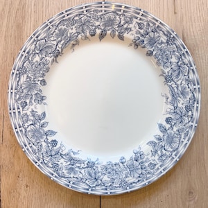 May include: A white ceramic dinner plate with a blue floral pattern around the rim. The pattern features a repeating design of flowers, leaves, and berries.