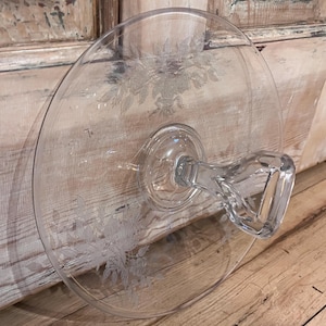 May include: Clear glass cake stand with a floral etched design. The stand features a round base and a handle for easy carrying. The etched floral pattern adds a touch of elegance to the serving piece.