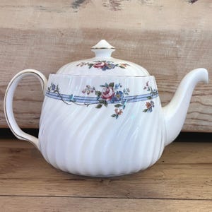 May include: White porcelain teapot with a spiral design and floral pattern. The teapot features a curved handle and spout, with a lid topped with a gold accent. A blue ribbon design with flowers encircles the teapot.