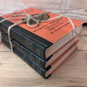 May include: A stack of three vintage hardcover books with orange covers and black spines. The top book is titled "Twenty Thousand Leagues Under The Sea" by Jules Verne.