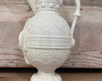 May include: Cream-colored ceramic pitcher with a spout and ornate handle. The pitcher features a detailed, embossed design with floral and geometric patterns. A horizontal band with text is visible on the body. The pitcher sits on a decorative base.