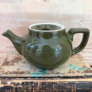 May include: A dark green ceramic teapot with a white rim and handle. The teapot is sitting on top of a stack of old books. The teapot has a spout and a lid with a small opening.