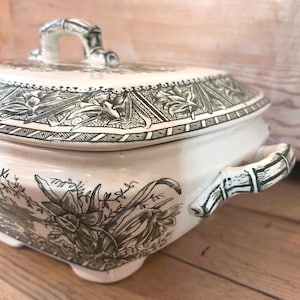 May include: Cream-colored ceramic serving dish with a lid, featuring a detailed green floral and botanical design. The dish has a square shape with rounded corners, a bamboo-style handle, and a matching lid.