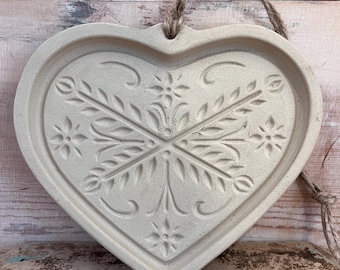 May include: A heart-shaped, off-white ceramic wall hanging with a floral and leaf design. The design is embossed into the ceramic. A natural rope is attached for hanging. The piece has a rustic, farmhouse aesthetic.