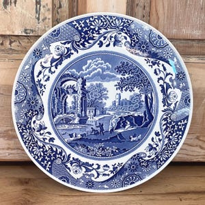 May include: A round, white and blue decorative plate with a detailed landscape scene in the center. The plate features a blue and white floral border and a classic, vintage design. The plate is approximately 10 inches in diameter.