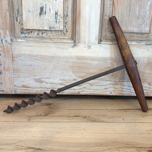 May include: An antique auger drill with a dark wooden handle and a spiral bit. The metal bit is a dark, rusty color. The handle is a T-shape. The tool is lying on a wooden surface.