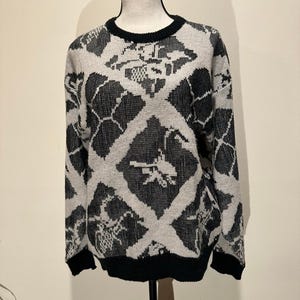 Puede incluir: Un suéter vintage con un patrón geométrico en blanco y negro. El suéter tiene cuello redondo y mangas largas con puños y dobladillo negros. El patrón es una forma de diamante repetida con diseños abstractos en su interior.