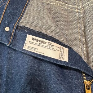 May include: Close-up of a pair of blue denim Wrangler jeans. The label reads "Wrangler" and includes the size 38 x 30. The jeans have a zipper and a button closure. The fabric is 100% cotton.