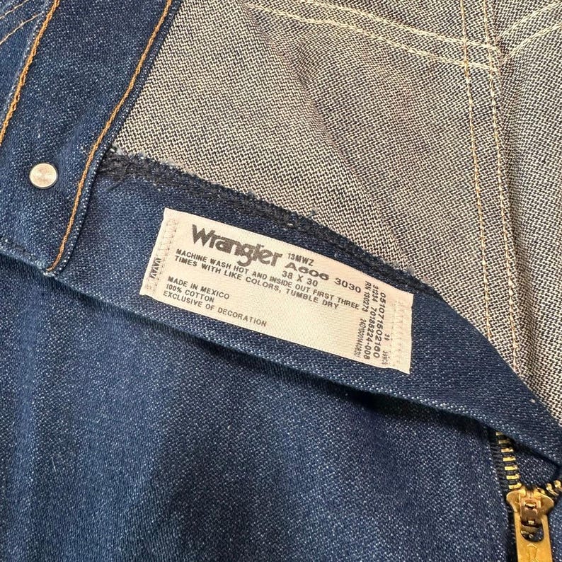 May include: Close-up of a pair of blue denim Wrangler jeans. The label reads "Wrangler" and includes the size 38 x 30. The jeans have a zipper and a button closure. The fabric is 100% cotton.