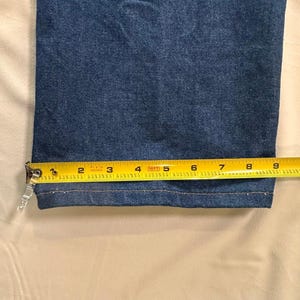 May include: Close-up of the hem of blue denim jeans. A yellow measuring tape shows the width of the hem is approximately 9 inches. The jeans are a medium wash denim.