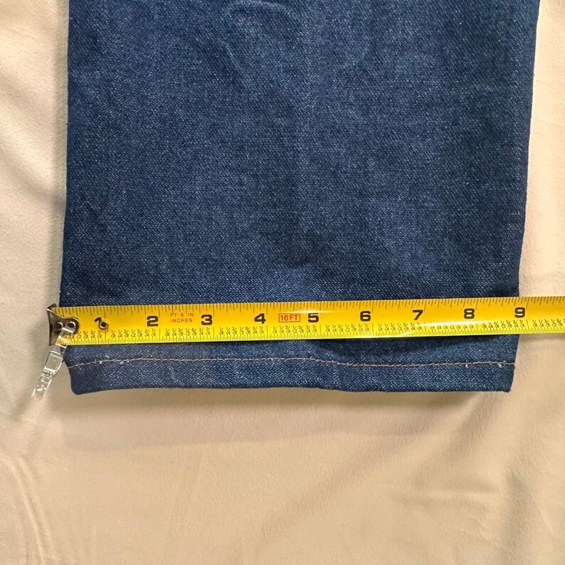 May include: Close-up of the hem of blue denim jeans. A yellow measuring tape shows the width of the hem is approximately 9 inches. The jeans are a medium wash denim.