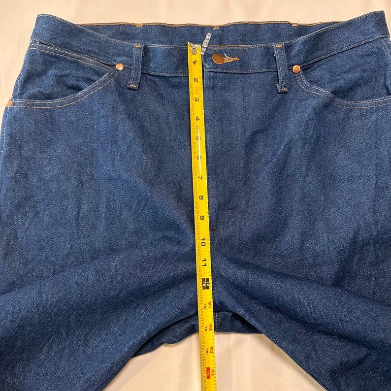 May include: Dark blue denim jeans with a straight leg design. A yellow measuring tape is placed vertically along the front of the jeans, showing a measurement of approximately 11 inches. The jeans have classic five-pocket styling and copper rivets.