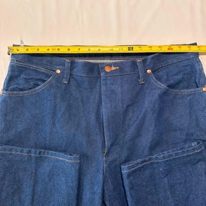 May include: Close-up of blue denim jeans with a button closure and copper rivets. A yellow measuring tape is laid across the waist of the jeans, showing the waist measurement in inches. The jeans have front and back pockets.