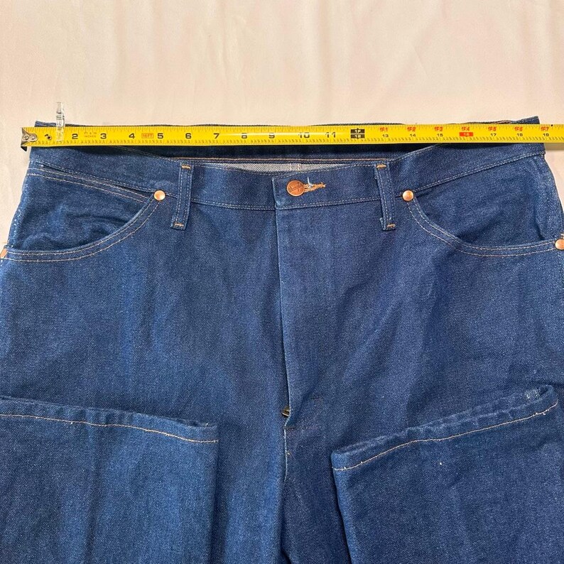May include: Close-up of blue denim jeans with a button closure and copper rivets. A yellow measuring tape is laid across the waist of the jeans, showing the waist measurement in inches. The jeans have front and back pockets.