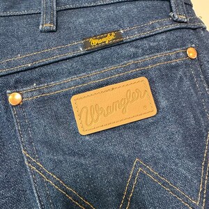 May include: Close-up of the back pocket area of blue denim Wrangler jeans. The jeans feature a brown leather patch with the Wrangler logo, gold stitching, and copper rivets. The Wrangler logo is also on a small black tag.