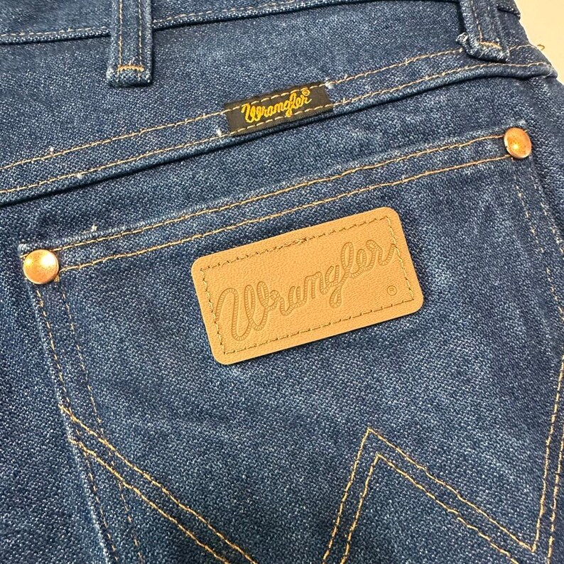 May include: Close-up of the back pocket area of blue denim Wrangler jeans. The jeans feature a brown leather patch with the Wrangler logo, gold stitching, and copper rivets. The Wrangler logo is also on a small black tag.