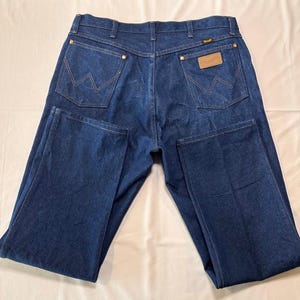 May include: Dark blue denim jeans with a classic five-pocket design. Features include copper rivets, a leather patch with the brand name, and the iconic 'W' stitching on the back pockets. The jeans are laid flat, showcasing the back view.