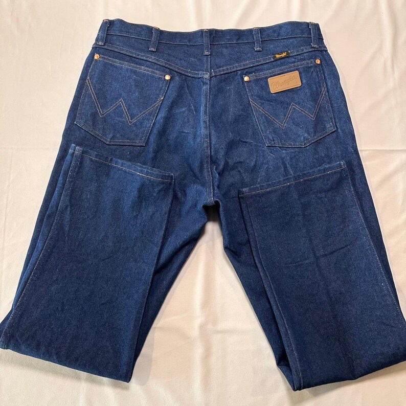 May include: Dark blue denim jeans with a classic five-pocket design. Features include copper rivets, a leather patch with the brand name, and the iconic 'W' stitching on the back pockets. The jeans are laid flat, showcasing the back view.