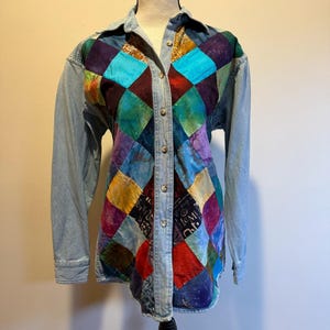 Può includere: Una camicia di jeans con maniche lunghe e un design patchwork. Il patchwork presenta un motivo a rombi di vari colori, tra cui blu, viola, rosso e oro. La camicia ha un colletto e chiusure con bottoni.