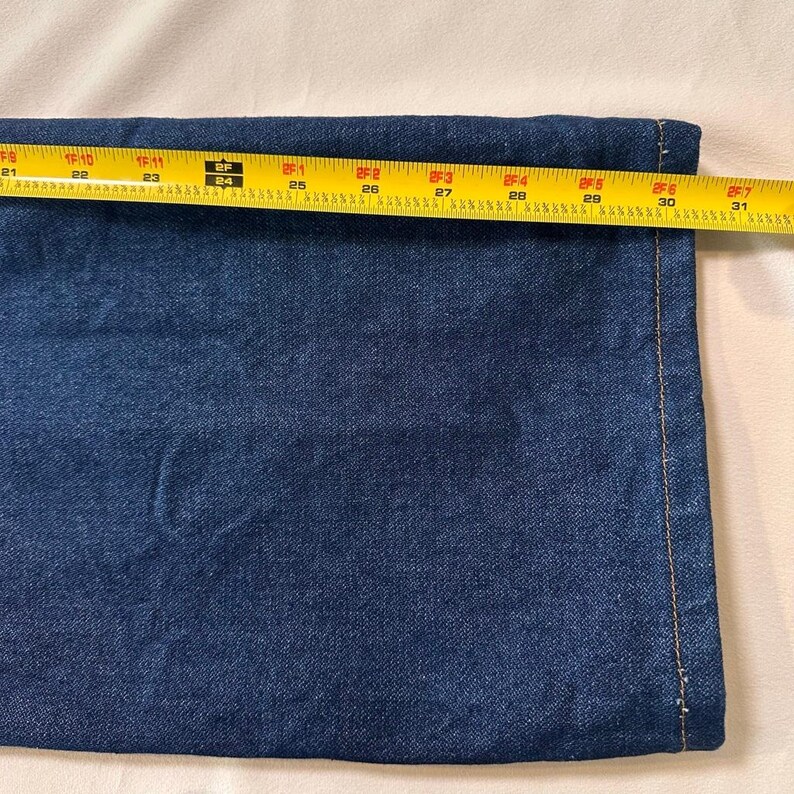May include: Close-up of a section of dark blue denim fabric. A yellow measuring tape is laid across the fabric, showing a measurement of approximately 30 inches. The fabric has a visible seam with tan stitching.
