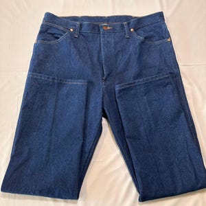 May include: Dark blue denim jeans with a classic five-pocket design. The jeans have a button and zipper closure, and copper-colored rivets. The fabric appears to be a medium-weight denim.