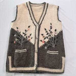 Puede incluir: Un chaleco sin mangas de punto crema y gris con cuello en V y cierre de botones. El chaleco tiene dos bolsillos y está decorado con bordados florales. El ribete y la tapeta de los botones son de color crema.