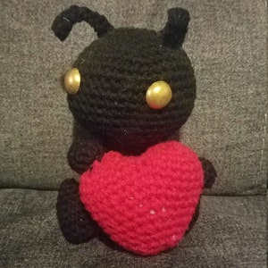 May include: A black crocheted plush toy with gold eyes holding a red crocheted heart.
