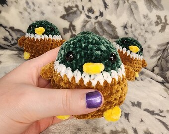 May include: Three crocheted plush ducklings. The ducklings are brown, green, and white with yellow beaks and black eyes.