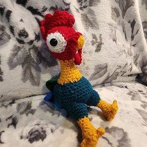 May include: A crocheted rooster toy with red, yellow, blue, and green yarn. The rooster has a large, white eye and a yellow beak.
