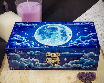 Puede incluir: Una caja de madera azul con un diseño pintado de luna y estrellas. La caja tiene un cierre dorado y está cerrada. La caja está sobre una superficie de madera.