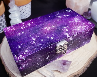 Puede incluir: Una pequeña caja de madera con un diseño de galaxia. La caja está pintada con un patrón de cielo estrellado morado y blanco y tiene un cierre plateado.