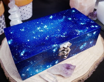 Puede incluir: Una caja de madera azul con un diseño de galaxia y un cierre plateado. La caja está sentada sobre una superficie de madera con un cristal morado y un cristal blanco.