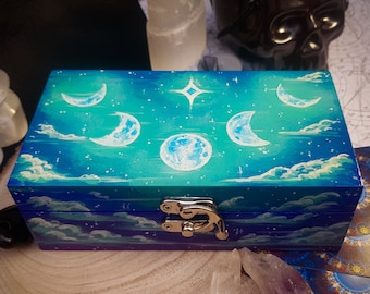 Può includere: Una scatola di legno con un cielo blu e viola dipinto sul coperchio. Il cielo è pieno di nuvole bianche e stelle. Ci sono cinque lune crescenti dipinte sul cielo, con una luna piena più grande al centro. La scatola ha una chiusura argentata.