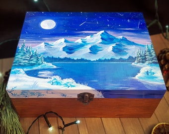 Puede incluir: Una caja rectangular de madera con una tapa pintada a mano que representa un paisaje de montaña nevado por la noche. La escena incluye una luna llena, un cielo azul oscuro estrellado, un lago helado y árboles de hoja perenne cubiertos de nieve.